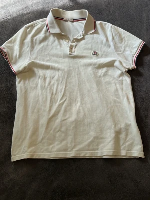 Vit Moncler pikétröja - Säljer en klassisk vit pikétröja från Moncler med röd och marinblå rand på krage och ärmslut. Tröjan har knappslå framtill och Moncler-logga på bröstet. Perfekt till jeans eller shorts för en clean och stilren look.