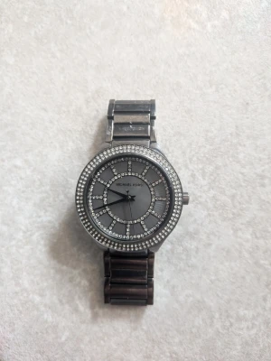 Silverfärgad klocka från Michael Kors - Stilren klocka från Michael Kors i silverfärg med glittrande stenar runt urtavlan och på urtavlans mönster. Klockan har en metallänk och klassisk rund form. Perfekt accessoar för dig som gillar lite extra bling. Ny pris 2500kr