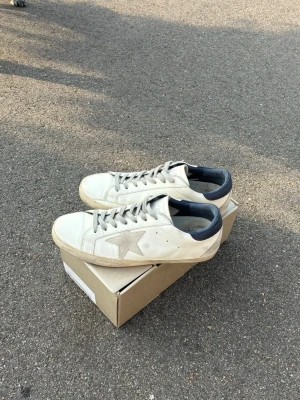 Riktigt feta golden goose ÄKTA! - Snygga vita sneakers från Golden Goose med den ikoniska stjärnan på sidan och mörkblå detaljer vid hälen. Skorna har en klassisk låg profil och är tillverkade i läder med slitna detaljer för en cool, avslappnad look. Perfekta för dig som gillar streetstyle.