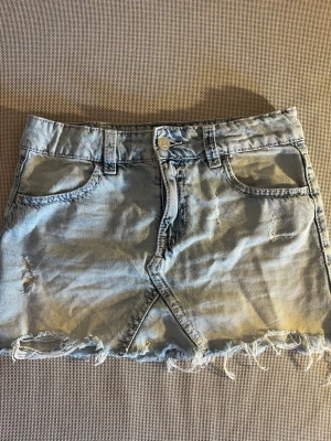 Lågmidjad ljusblå jeanskjol med fransig kant - Säljer en ljusblå jeanskjol som är lågmidjad och har en  fransig nederkant och slitna detaljer. Kjolen har fem fickor, bälteshällor och en snygg omlott-effekt framtill. Bakfickorna har broderade detaljer. Perfekt för dig som gillar en trendig stil.
