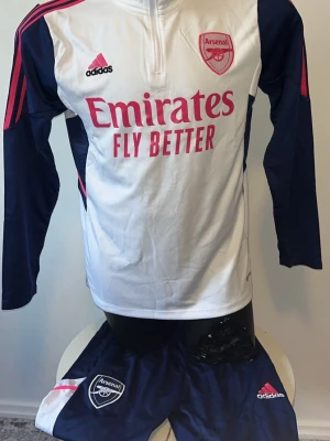 Arsenal träningsset från Adidas - Snyggt Arsenal träningsset från Adidas med långärmad vit tröja och marinblå ärmar, samt matchande marinblå byxor. Rosa detaljer och loggor på både tröja och byxor. Perfekt för fotbollsträning eller chill. Materialet är lätt och andas.