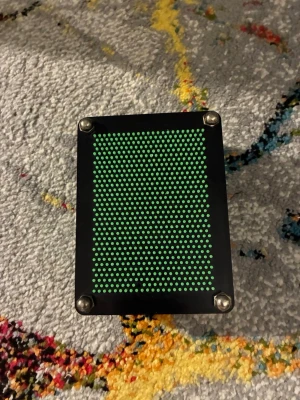 Grön pin art leksak - En klassisk pin art leksak med gröna plastpinnar och svart ram. Tryck handen eller andra föremål mot pinnarna för att skapa 3D-former som syns på andra sidan. Perfekt som dekoration eller för att leka med olika mönster.