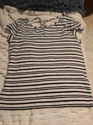 Randig vit och svart topp med knyt - Säljer en kortärmad topp i vitt med svarta ränder och ribbad struktur. Toppen har en v-ringning med snygg knyt-detalj i nacken och är i en mjuk, stretchig kvalitet. Perfekt till jeans eller kjol för en chill look.