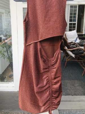 kort festklänning med cut out - Säljer en glittrig guld/brun kortklänning med snygg cut out-detalj i midjan och dragsko framtill. Klänningen är ärmlös och har en unik design som verkligen sticker ut. Perfekt för dig som vill ha något extra till nästa fest. Klänningen är från HM och har bara använts vid ett tillfälle 