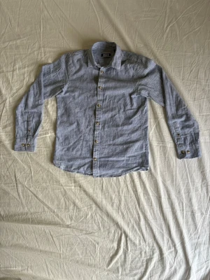 Blå och vit-randig skjorta från Jack & Jones - Säljer en klassisk långärmad skjorta från Jack & Jones i storlek 152. Skjortan är blå- och vitrandig med knappar framtill och på ärmsluten. Tillverkad i mjuk bomull/linne, perfekt för en clean och stilren look.