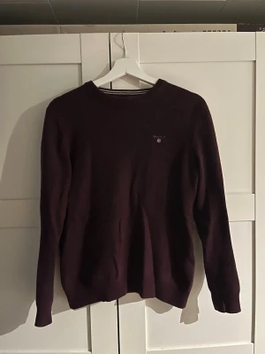 Mörklila/ röd stickad tröja från Gant - Snygg mörklila/röd stickad tröja från Gant med rund halsringning och diskret logga broderad på bröstet. Tröjan har långa ärmar och ribbade muddar vid ärmslut och nederkant. Storlek M men sitter som S/XS Perfekt för dig som gillar klassisk och stilren design. Riktigt skönt och strechigt material. Pris kan diskuteras. Hör av er vid frågor!