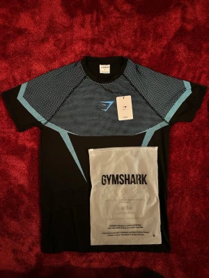 Svart och blå Gymshark träningströja - Snygg svart träningströja från Gymshark med blått och vitt grafiskt mönster över bröst och axlar. T-shirten har korta ärmar och är tillverkad i ett lätt och stretchigt funktionsmaterial som passar perfekt till gymmet eller löpning. 