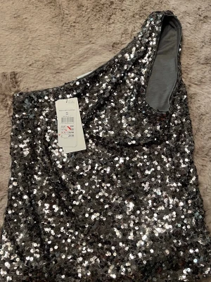 Glittrig oneshoulder topp i silver - Säljer en superglittrig oneshoulder topp i silver med massor av paljetter. Perfekt för dig som vill sticka ut och glänsa. Toppen är axelbandslös på ena sidan och har en snygg, figurnära passform.