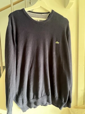 Mörkblå stickad tröja från Lacoste - Mörkblå stickad tröja från Lacoste med rund halsringning och den klassiska gröna krokodilloggan på bröstet. Tröjan har ribbade muddar vid ärmslut och nederkant. Perfekt för dig som gillar stilrena och tidlösa plagg. Storlek XL