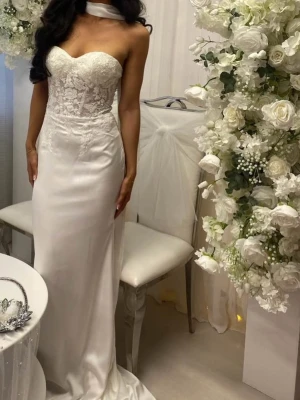 Vit lång spetsklänning med släp - Elegant vit långklänning med figurnära passform och hjärtformad urringning. Överdelen har vacker spets och broderade detaljer, medan kjolen är slät och faller mjukt ner mot golvet. Klänningen är axelbandslös och har ett litet släp för extra effekt.