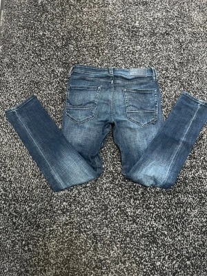 slim/glenn jack & jones - Säljer ett par mörkblå slim fit jeans med snygga slitningar och klassiska bakfickor. Jeansen har en bra passform och är tillverkade i denim med lätt stretch för extra komfort. Perfekta till en avslappnad och trendig stil.