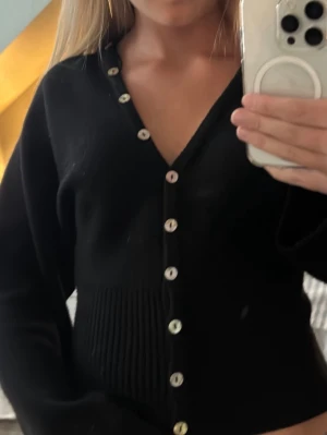 Svart ribbad kofta från Gina Tricot - Snygg svart stickad kofta från Gina Tricot med v-ringning och pärlemorskimrande knappar framtill. Koftan har ribbad midja som ger en smickrande passform och långa ärmar. Perfekt att styla till jeans eller kjol. Extra knapp kommer med, skriv om ni har frågor❣️