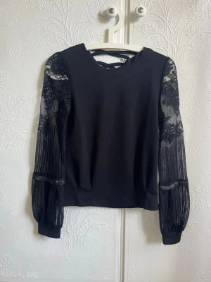 Svart topp med spetsärmar - Snygg svart topp med transparenta spetsärmar och diskreta blommönster. Ärmarna är långa och har en lätt puffig form med ribbade muddar. Toppen har en enkel rund halsringning. Perfekt för dig som vill ha något extra till outfiten.