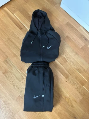 Nike tracksuit  - Snygg svart hoodie från Nike med dragkedja framtill och justerbar huva med snören. Hoodien har ribbade muddar vid ärmslut och nederkant samt två fickor. Ikonisk Nike-logga på bröstet. Perfekt för en sportig och avslappnad stil.