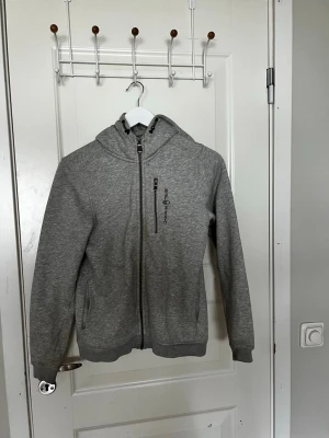 Grå hoodie från Sail Racing - Säljer en grå hoodie från Sail Racing med dragkedja framtill, två sidofickor och en bröstficka med dragkedja. Hoodien har justerbar huva med snören och mjukt fleecefoder på insidan. Perfekt för chill dagar eller när du vill ha något bekvämt och stilrent.