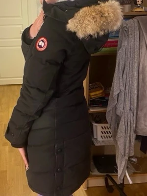 Svart lång dunjacka med pälskant - Säljer en svart lång dunjacka från Canada Goose med huva och beige pälskant. Jackan har en klassisk patch på ärmen, knappar vid ärmslut och längs sidorna samt dragkedja framtill. Perfekt för kalla vinterdagar.