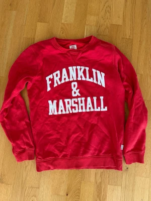 Röd sweatshirt från Franklin & Marshall - Cool röd sweatshirt från Franklin & Marshall med vit logga tryckt framtill. Klassisk rund halsringning och ribbade muddar vid ärmslut och nederkant. Perfekt för en avslappnad och sportig stil.