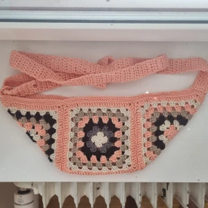Virkad magväska i mormorsrutor  - Unik handgjord magväska virkad i mormorsrutor. Rosa botten, med beige, svart och grått. Justerbart band så den kan bäras crossbody eller runt midjan. Perfekt för dig som vill ha en färgglad och retro accessoar till din outfit. 100% bomull och stärkt för att hålla formen och vara extra slitstark. Stängs med tryckknapp.