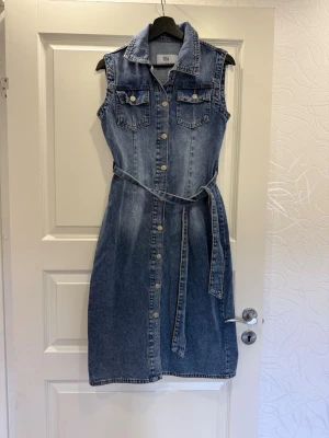 Ärmlös jeansklänning med bälte - Snygg ärmlös jeansklänning i blå denim med knappar hela vägen framtill, krage och två bröstfickor. Medföljande bälte i samma material markerar midjan. 