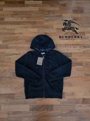 Svart Burberry Hoodie - Letar du efter en stilren och lyxig hoodie? Denna helt nya grå Burberry-hoodie kombinerar elegans med hög komfort. Den har Burberrys ikoniska rutmönster i fodret och en diskret logga på bröstet för en exklusiv touch. Tillverkad av mjukt, slitstarkt material och utrustad med dragkedja samt huva – både praktisk och stilfull.  📌 Skick: Ny med taggar. 📦 Snabb leverans – skickas samma dag (om ej hinder)
