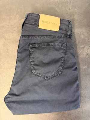 Mörkblå chinos från Rose & Born, stl 33/34 - Snygga mörkblå chinos från Rose & Born i smal passform. Byxorna har klassiska fickor fram och bak, bälteshällor och stängs med knapp och dragkedja. Tillverkade i mjukt bomullstyg som ger en skön känsla och stilren look.