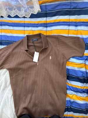 Brun ribbad topp från Polo Ralph Lauren - Snygg brun ribbad topp från Polo Ralph Lauren med korta ärmar och liten dragkedja vid halsen. Tillverkad i mjuk bomull och har en diskret logga broderad på bröstet. Perfekt för dig som gillar klassisk och stilren design.