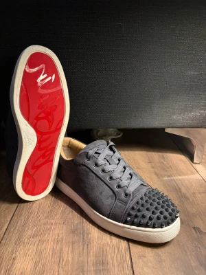 Christian loubutin skor  - Unika svarta sneakers med mattsvarta nitar på tån och ikonisk röd sula. Skorna har svarta skosnören, vit yttersula och är tillverkade i mockaliknande material. Perfekt för dig som vill sticka ut med en edgy och trendig look.