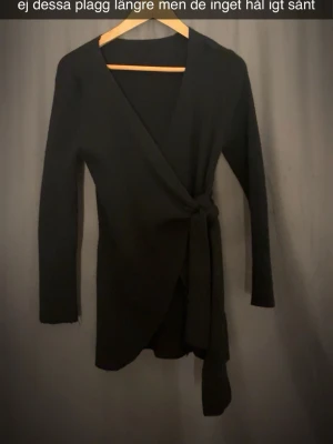Svart omlottkofta med knytband - Stilren svart omlottkofta med långa ärmar och knytband i midjan. Koftan har en smickrande V-ringning och är gjord i ett mjukt, stretchigt material. Perfekt att slänga över en topp för en enkel men snygg look.