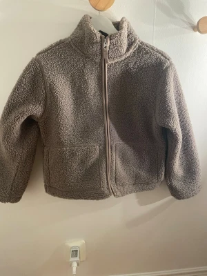 Brun teddyjacka med dragkedja - Mysig brun teddyjacka med hög krage och hel dragkedja framtill. Jackan har två stora fickor fram och är gjord i fluffigt, mjukt teddy-material. Perfekt jacka att styla Jeans eller mjukisbyxor till 