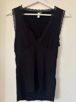 Svart spetskantad linnetopp - Snygg svart linnetopp med v-ringning och spetsdetaljer längs urringning och axelband. Toppen är ärmlös och har en feminin, enkel design som passar perfekt till många olika outfits.