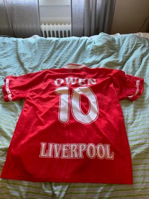 Liverpool fotbollströja Owen #10 - Röd Liverpool fotbollströja med vita detaljer och tryck. På ryggen står det 'Owen' och nummer 10. Framtill finns Liverpool FC:s klubbmärke och Carlsberg-logga. Tröjan har korta ärmar och klassisk krage. Perfekt för dig som älskar fotboll och vill visa ditt stöd för laget.