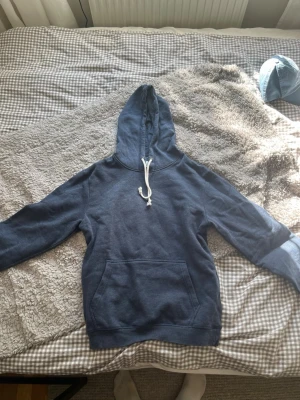 Blå hoodie med ficka och dragsko - Mysig blå hoodie med stor känguruficka framtill och vit dragsko i huvan. Hoodien har långa ärmar och ribbade muddar vid ärmslut och nederkant. Perfekt för chill dagar eller när du vill ha något bekvämt och enkelt.