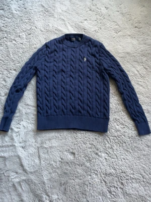 Mörkblå kabelstickad tröja - Snygg mörkblå kabelstickad tröja med rund halsringning och ribbade muddar vid ärmslut och nederkant. Tröjan har ett diskret broderat emblem på bröstet och är perfekt för dig som gillar klassisk stil med en twist.