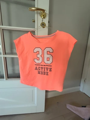 Neonorange t-shirt med tryck - Cool neonorange t-shirt med stort tryck av siffran 36 och texten 'Active Mode' på framsidan. T-shirten har korta ärmar och en liten off shoulder. Tröjan e ifrån hm tror jag! 