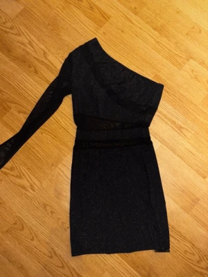 Svart glittrig one shoulder klänning - Svart glittrig kortklänning från SHEIN SXY med en ärm och one shoulder-design. Klänningen har mesh-detaljer vid midjan och överdelen, vilket ger en extra snygg touch. Perfekt för dig som vill sticka ut med en trendig look.