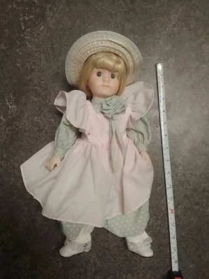 Porslinsdocka med ljusrosa klänning - Söt porslinsdocka med blont hår, stora bruna ögon och en ljusrosa klänning med puffärmar och stor rosett. Dockan har gröna byxor med vita prickar, vita skor och en matchande ljusgrön hatt. Perfekt som dekoration eller samlarobjekt.