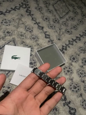 Silverfärgat armband från Lacoste - Snyggt armband i silverfärgad metall från Lacoste. Armbandet har en stilren, länkad design med diskreta detaljer och Lacoste-loggan ingraverad på spännet. Perfekt accessoar för dig som gillar en clean och modern look.