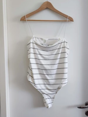 Vit body med svarta ränder - Säljer en vit body med tunna svarta horisontella ränder och smala axelband. Bodyn har en enkel och stilren design med tryckknappar i grenen. Tillverkad i mjukt och stretchigt material, perfekt för en avslappnad och trendig look.