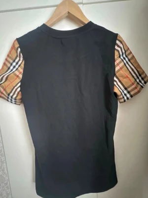 Svart Burberry t-shirt med rutiga ärmar - Snygg svart t-shirt från Burberry med klassiska beige-rutiga korta ärmar. T-shirten har rund halsringning och är gjord i mjuk bomull. Perfekt för dig som vill ha en stilren look med en twist av Burberrys ikoniska mönster. Ny utan prislapp