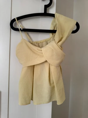Gul axelbandslös topp med rosett - Trendig gul topp i strukturerat tyg med stor rosettdetalj framtill. Toppen är axelbandslös på ena sidan och har ett smalt axelband på andra, vilket ger en asymmetrisk och modern look. Perfekt för dig som vill sticka ut i sommar.