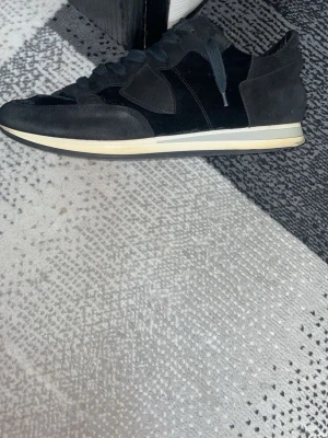 Svarta sneakers i mocka och textil - Snygga svarta sneakers med ovandel i mocka och textil, samt vita sulor med retrovibbar. Skorna har svarta skosnören och en stilren design med diskreta paneler och detaljer. Perfekta för dig som gillar en clean och klassisk look.