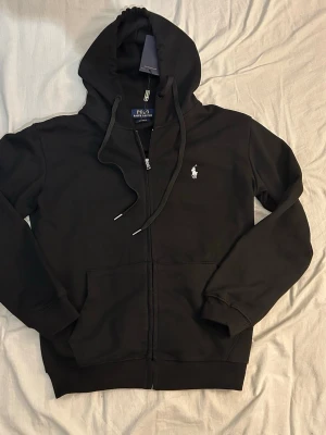 Svart hoodie från Polo Ralph Lauren - Snygg svart hoodie från Polo Ralph Lauren med dragkedja framtill, justerbar huva och fickor på sidorna. Klassisk vit broderad logga på bröstet. Tillverkad i mjukt bomullsmaterial som är skönt att bära. Perfekt för en avslappnad och stilren look.