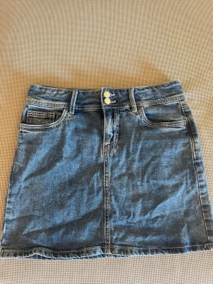 Blå jeanskjol med dubbla knappar - Snygg blå jeanskjol i klassisk modell med dubbla guldfärgade knappar framtill och femficksdesign. Kjolen har bälteshällor och är tillverkad i ett slitstarkt denimtyg. Perfekt för dig som gillar en enkel men trendig look.