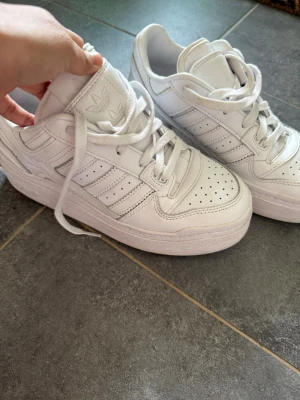 Vita Adidas sneakers med platåsula - Säljer ett par helvita Adidas sneakers med klassiska tre ränder på sidan och platåsula. Skorna är låga och har perforerad tå för ventilation samt Adidas-logga på plösen och sidan. Tillverkade i läder med vita skosnören för en clean look.