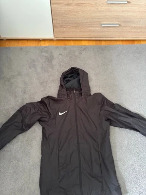 Svart Nike vindjacka med huva - Snygg svart vindjacka från Nike med huva och dragkedja framtill. Vid funderingar eller frågor är det bara att höra av sig!