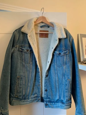 Levi's blå fodrad jeansjacka M - Klassisk blå jeansjacka från Levi's i storlek M med vit teddyfoder på insidan och kragen. Jackan har två bröstfickor med lock och knappar samt sidofickor. Perfekt för dig som vill ha en tidlös och snygg look med extra värme.