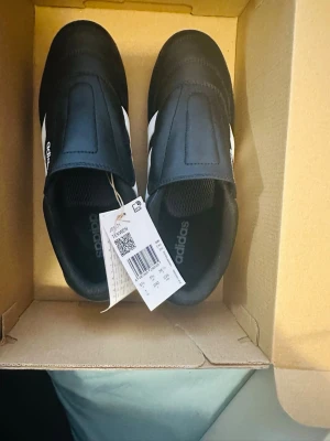 Svarta Adidas sneakers med vita ränder - Snygga svarta sneakers från Adidas med klassiska vita ränder på sidan och diskret logga på plösen. Skorna har en låg profil och är tillverkade i syntetmaterial med textilfoder. Perfekta för en clean och sportig look.