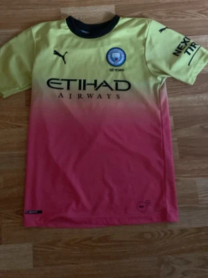 Manchester City matchtröja  - Säljer en Manchester City matchtröja från Puma i en snygg gul till rosa färgövergång. Tröjan har svart krage, klubbmärke på bröstet och Etihad Airways-tryck framtill. Perfekt för fotbollsträning eller att bära till vardags.