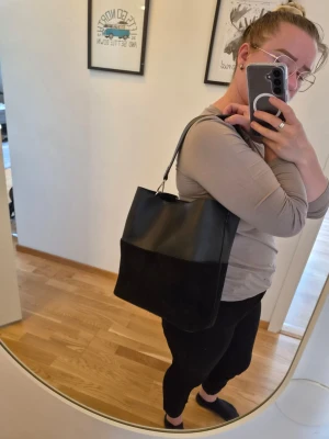 Svart axelväska i skinn och mocka. Läs innan köp! - Snygg svart axelväska med bred axelrem. Väskan har en kombination av skinn imitation upptill och mocka imitation nedtill. Rymlig modell, enkel och stilren design som passar till det mesta. Den har ett löstagbart fack som sitter i mitten av väskan. Och kan förekomma små fläckar som knappt syns. Botten är lite skräpig men går att rengöra. Har  funnits ett längre axelband också men inte längre. Nypris 600 kr OBS katt finns i hemmet