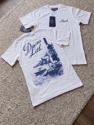 Vit t-shirt med blått tryck - Säljer en vit t-shirt med blått tryck på ryggen som föreställer en vinflaska, glas och naturmotiv samt texten 'Døss og Lith'. Framtill finns en liten blå logga med texten 'Marcel'. Klassisk passform och rund hals.